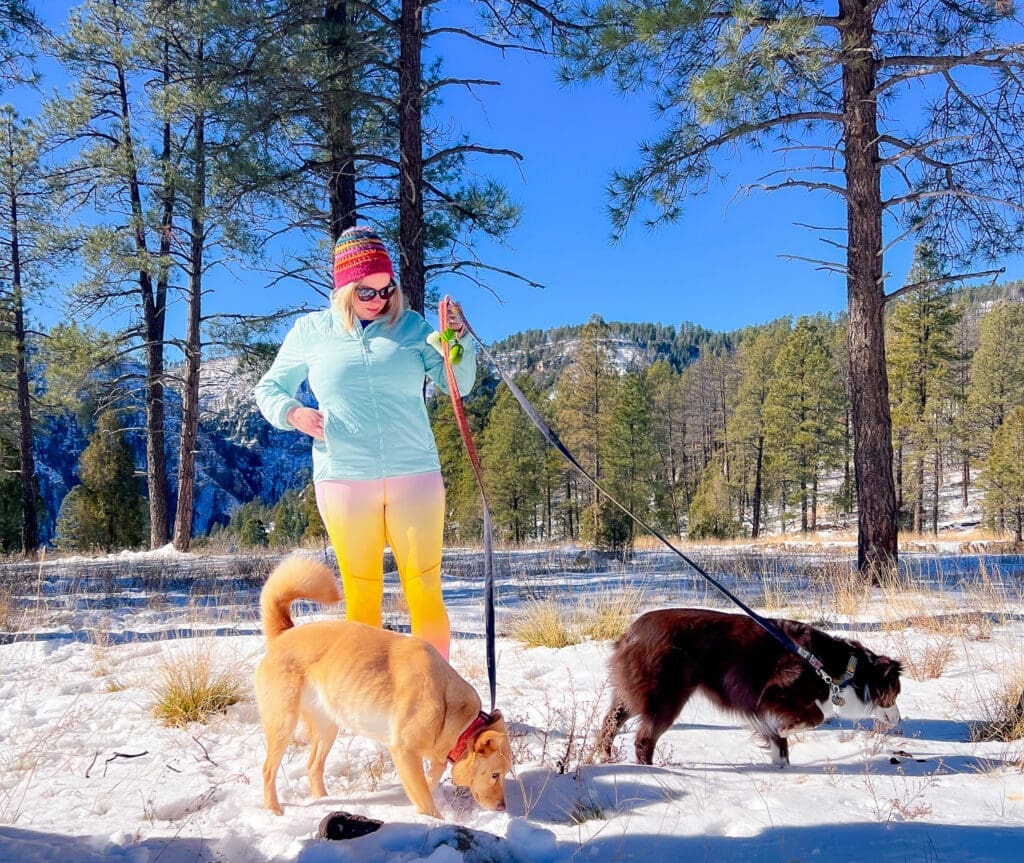 Blair stands in snow with June and Margot on leash. Mountains and trees are behind them.